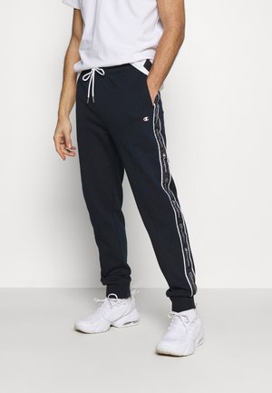 Mann trägt marineblaue Champion-Trainingshose mit weißem Logostreifen und weißen Sneakers, steht mit einer Hand in der Tasche.