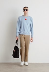 Svetlomodrá mikina s malou červenou nášivkou, béžové nohavice, biele tenisky a čierna taška. Okuliare dopĺňajú outfit.