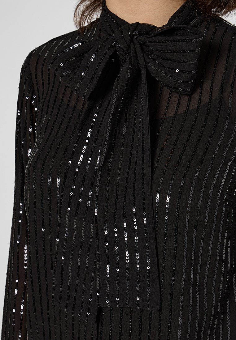 Zwarte blouse met verticale strepen van pailletten, een strik aan de hals en een doorschijnende stofstructuur.