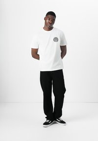 Hvid bomulds t-shirt med et rundt sort grafisk design på brystet, parret med løse sorte bukser og sorte sneakers med hvide striber.