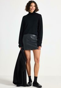 Sort turtleneck sweater med lange ærmer, parret med en tætsiddende sort læder mini nederdel. Modellen holder en sort frakke og bærer sorte sokker og sko.