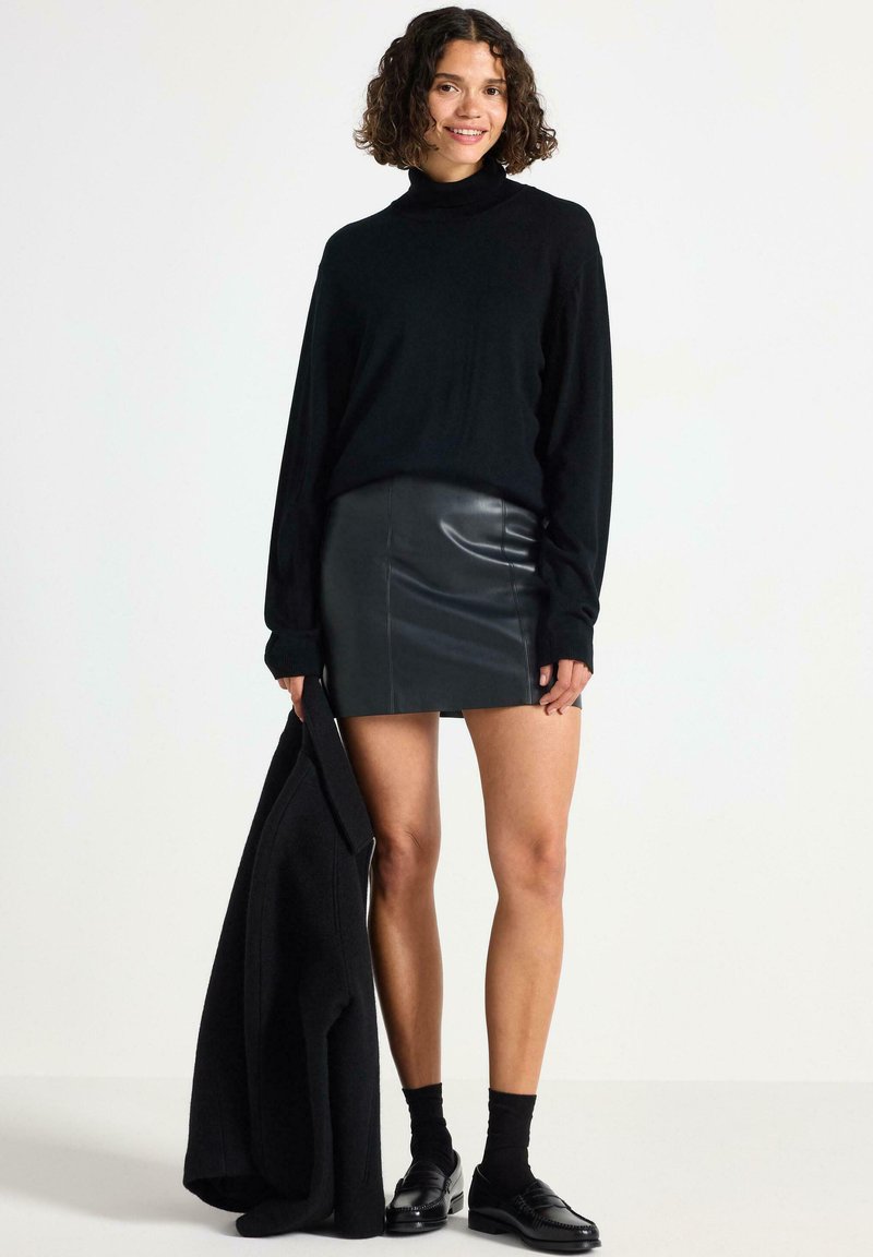 Sort turtleneck sweater med lange ærmer, parret med en tætsiddende sort læder mini nederdel. Modellen holder en sort frakke og bærer sorte sokker og sko.