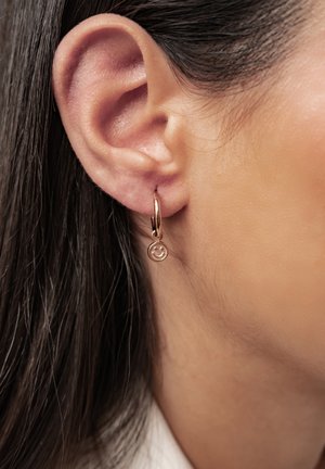 Primer plano de una oreja con cabello oscuro que lleva un pequeño aro de oro con un charm de cara sonriente.