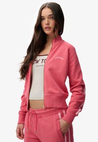 Sudadera rosa con cremallera y bajo acanalado, con detalles blancos y un logo. Combinada con pantalones a juego, mostrando un diseño casual y atlético.