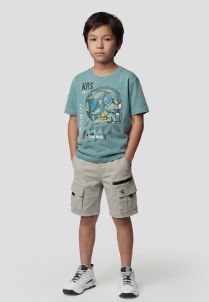 Niño de pie con las manos en los bolsillos, vistiendo una camiseta gráfica color teal con una calavera, pantalones cortos tipo cargo beige y zapatillas blancas sobre fondo gris.