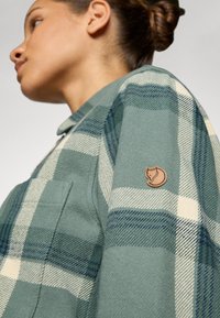 Donna con capelli raccolti in un bun che indossa una camicia a quadri verde e crema con una patch a forma di volpe sulla manica, che guarda verso sinistra su uno sfondo semplice.