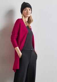 Magenta cardigan přes černý top, spárovaný s černými kalhotami. Cardigan má dlouhé rukávy a otevřený design vpředu.