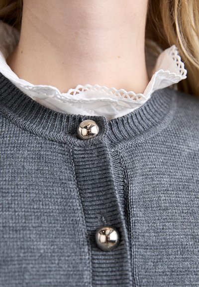 Harmaa neulecardigan, jossa on teksturoidun viimeistelyn, pyöreä pääntie, kaksi kiiltävää hopeista nappia ja valkoinen pitsikauluksellinen paita alla.