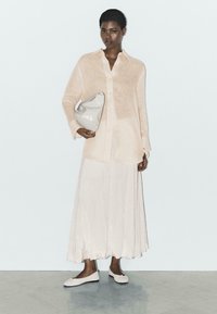 Frau trägt eine hellrosa transparente Bluse und einen fließenden Rock, hält eine weiße Clutch und steht auf einem Marmorboden vor einer schlichten weißen Wand.