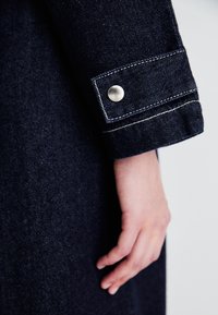 Dunkelblauer Denim-Stoff mit einem Bündchen, das über weiße Nähte und einen silbernen Druckknopf verfügt. Nahaufnahme einer Hand, die auf dem Denim ruht.