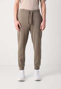 Muške jogger hlače od mekog, khaki materijala. Imaju pojas s vezicom, sužene nogavice i rebraste manžete. Uparene s bijelim tenisicama.