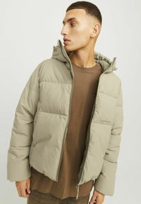 Veste kaki matelassée avec fermeture éclair et capuche, présentant une coupe ample et des panneaux cousus. Associée à un t-shirt marron à col rond en dessous.