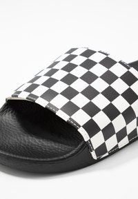 Sandale à glissière en damier noir et blanc avec une large bride, une tige texturée et une semelle noire lisse. Détails de surpiqûres le long des bords.