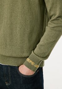 Suéter verde oliva con puños acanalados que muestra "Pepe Jeans" en amarillo. Textura suave y diseño ajustado. Combinado con jeans de mezclilla oscura.
