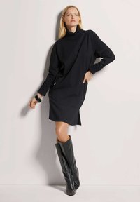 Schwarzes Strickkleid mit hohem Kragen, ausgestattet mit Reißverschlusstaschen und einem lässigen Schnitt. Kombiniert mit hohen schwarzen Lederstiefeln.