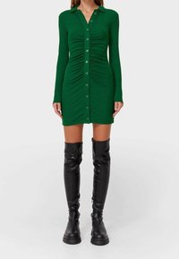 Robe verte à manches longues avec un col à revers, des détails froncés et des boutons sur le devant. Portée avec des bottes noires montantes. Texture lisse.