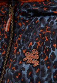 Vadderad jacka med ett mönster i blå och svart leopardprint och orangea accenter. En broderad lapp med texten "Gör din grej" är synlig på framsidan.