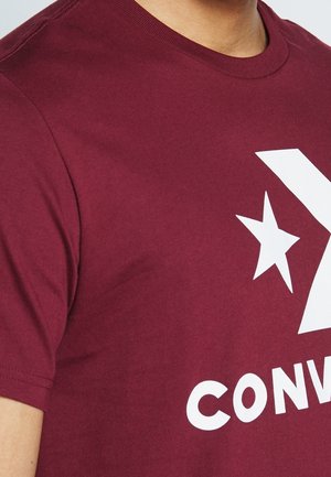 Weinrotes kurzärmliges Hemd mit weißem Converse-Stern und teilweise sichtbarem Logo am oberen Oberkörper einer Person.