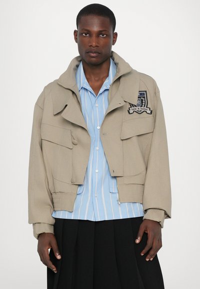 Beige cropped Jacke mit übergroßer Kapuze, mehreren vorderen Taschen, gerippten Manschetten und besticktem Patch. Darunter ein blau gestreiftes Hemd.