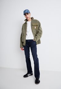 Giacca in pile verde oliva con una tasca marrone, maglietta bianca a girocollo, jeans scuri e sneakers nere con strisce bianche, abbinati a un cappellino di denim.
