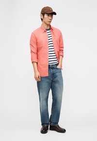 Muškarac koji nosi smeđu kapu, koraljnu košulju s dugim rukavima preko majice s tamnoplavim i bijelim prugama, plave traperice i smeđe čizme, stoji s jednom rukom u džepu.