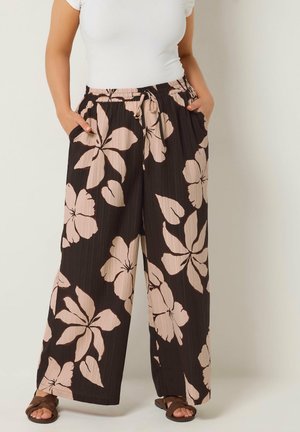 Kvinde iført sorte bukser med brede ben og stort beige blomsterprint, hvid kortærmet top og brune sandaler, med hænderne i lommerne, stående.