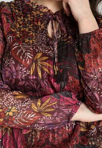 Blusa trasparente a fiori in un profondo bordeaux, caratterizzata da un colletto con laccetto, maniche morbide e delicati polsini elasticizzati.