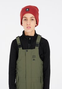 Salopette da sci verde con bretelle regolabili, chiusura con zip e tasche laterali, indossata sopra una giacca nera con zip, con un berretto rosso con logo.