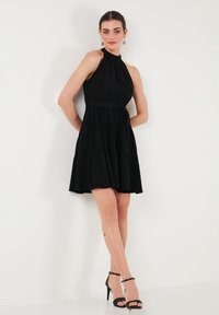 Robe noire à bretelles, tissu léger, design à volants, taille ajustée avec ceinture, longueur au-dessus du genou. Associée à des sandales à talons hauts noires.
