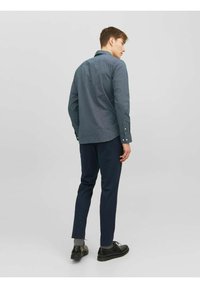 Camicia blu scuro a fantasia con colletto button-down, indossata con pantaloni neri e scarpe nere, vista da dietro. Texture sottile visibile.