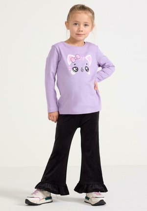 Camicia a maniche lunghe color lilla chiaro con grafica di un gatto, abbinata a pantaloni neri a zampa con orli arricciati. Scarpe da ginnastica con accenti di colore.