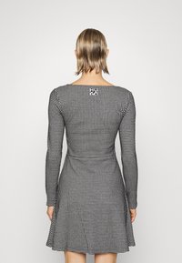 Schwarz-weißes Kleid mit Hahnentritt-Muster, langen Ärmeln, figurbetontem Oberteil und weitem Rock. Logo-Label gut sichtbar am Rücken des Ausschnitts.