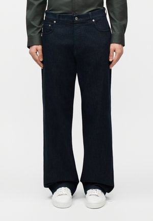 Mand iført mørke vidde bukser, mørkegrøn skjorte og hvide sneakers, stående mod en ensfarvet hvid baggrund.