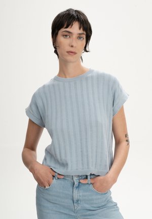 Personne aux cheveux courts et foncés portant un pull à manches courtes côtelé bleu clair et un jean en denim clair, les mains dans les poches.