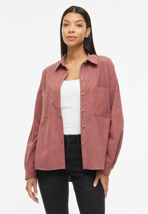 Femme portant une veste boutonnée en velours côtelé couleur rose poussiéreux sur un haut blanc et un pantalon noir, debout devant un fond blanc uni.