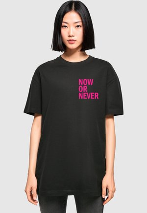 Sort bomulds t-shirt med korte ærmer, med pink trykt tekst "NOW OR NEVER" på forsiden i en stablet opstilling.