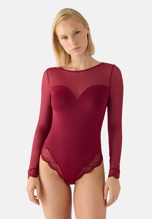 Mujer que lleva un body burdeos de manga larga con ribete de encaje y parte superior del pecho y mangas transparentes, de pie contra un fondo sencillo.