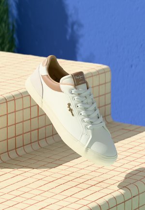 White leather sneaker with tan accents, round toe, and lace-up design. Features a logo on the side and textured rubber sole.
