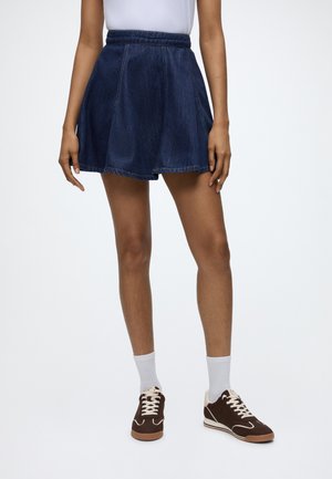 Personne portant une jupe en jean bleu foncé, des chaussettes blanches et des baskets marron foncé à lacets blancs, debout devant un fond uni.