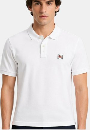 Uomo che indossa una polo bianca semplice a maniche corte con un piccolo logo a toppa sul petto sinistro.