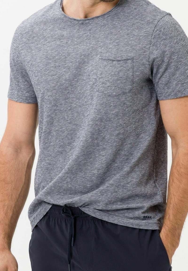 Homme portant un t-shirt gris chiné à manches courtes avec une poche poitrine et un pantalon noir à cordon, posant avec une main dans la poche.