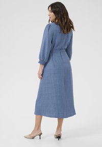 Robe longue bleue texturée avec des manches bouffantes et une ceinture à la taille ; longueur mi-mollet avec une coupe décontractée, associée à des sandales à talons nus.