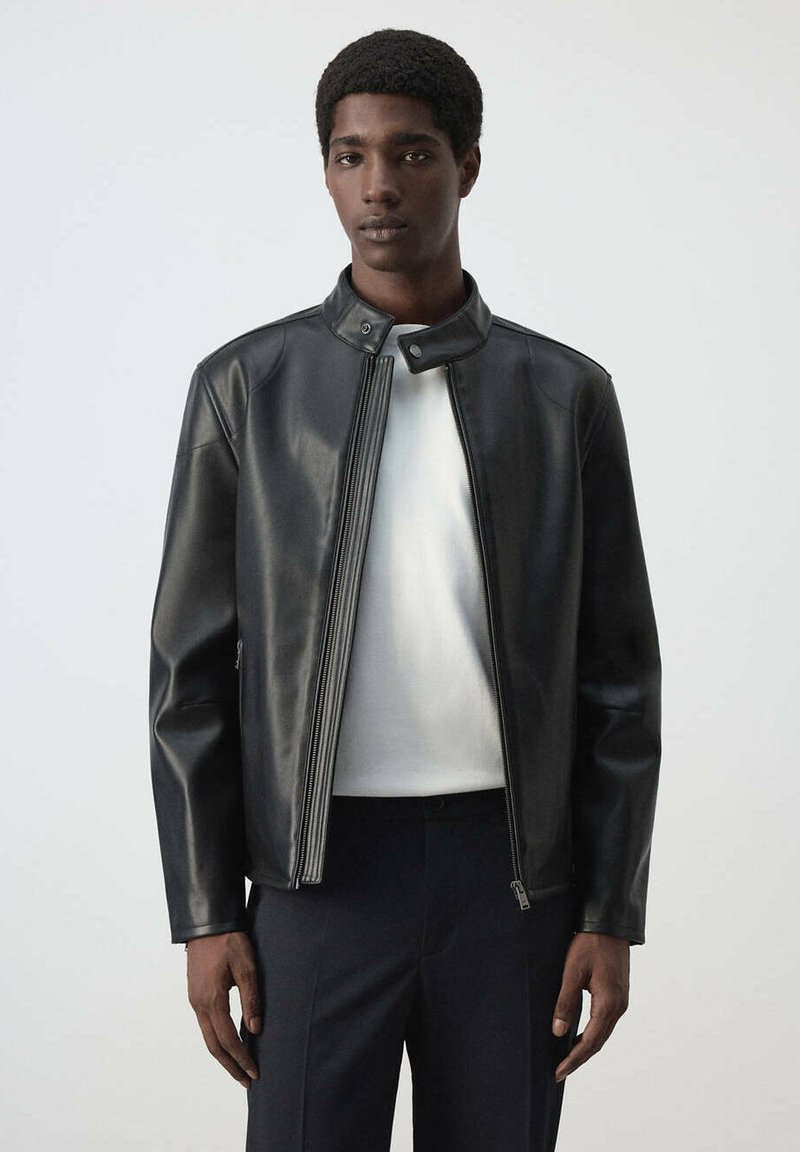 Jeune homme portant une veste en cuir noire sur une chemise blanche, debout devant un fond clair uni, regardant devant lui.