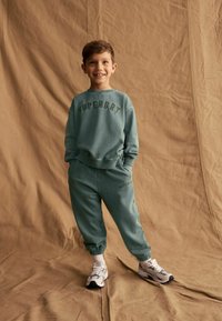 Tyrkysová bavlnená mikina a nohavice s logom "SUPERDRY" v tmavozelenej farbe, so vzorovanými manžetami a pásom; spárované so šedými a čiernymi teniskami.