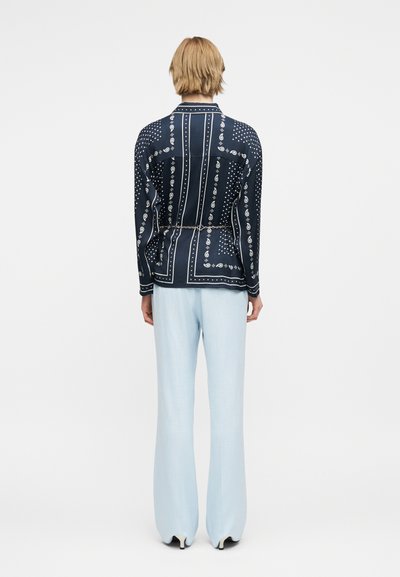 Άτομο με κοντά ξανθά μαλλιά που φοράει μαύρο πουκάμισο με σχέδια και μακριά μανίκια, ανοιχτό μπλε παντελόνι με φαρδιά πόδια και λευκά τακούνια, γυρισμένο από την πλάτη.