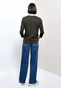 Olijfgroene shirt met lange mouwen gecombineerd met wijde blauwe jeans. Het shirt heeft een ronde hals; de jeans hebben achterzakken en een klassieke pasvorm.