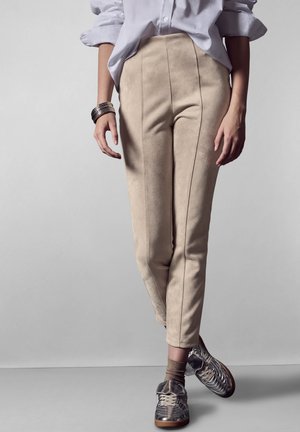 Pantalons en suède beige avec des détails de couture, associés à des baskets argent métallisé et des chaussettes brunes. La coupe est ajustée avec un design à jambe fuselée.