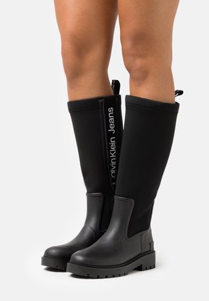 Bottes noires montantes au genou avec semelles épaisses portées par une personne, arborant le logo Calvin Klein Jeans sur le panneau latéral intérieur.