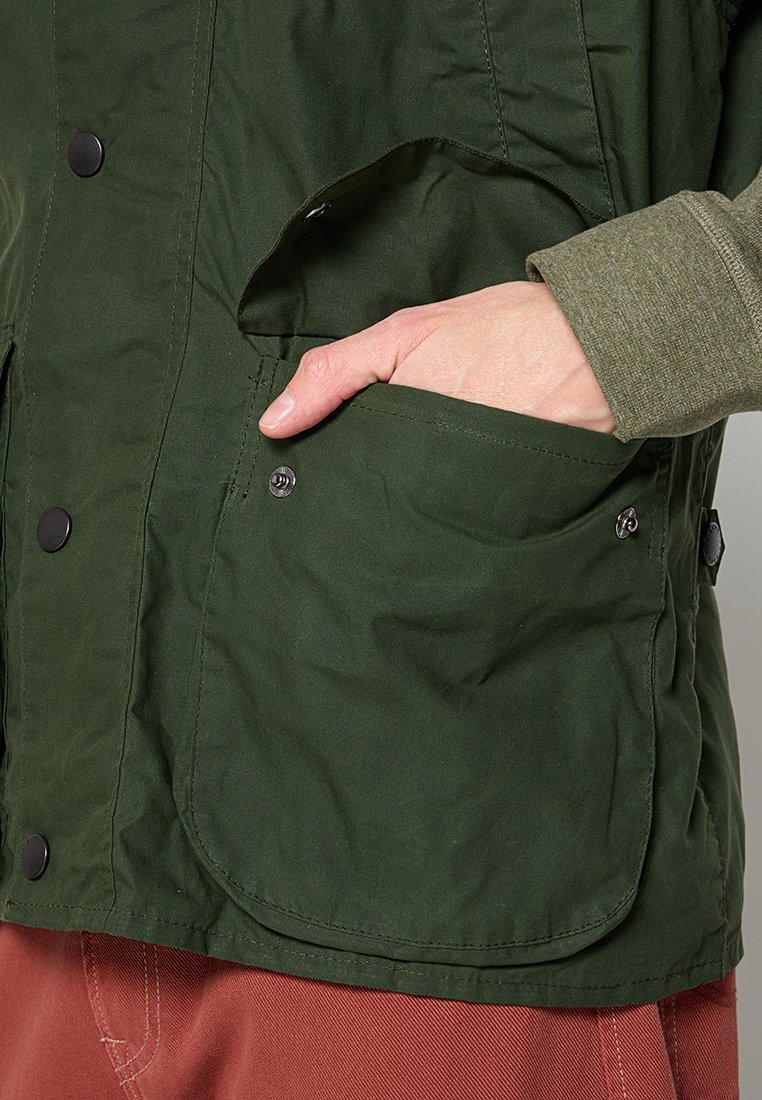 Eine Hand steckt in die vordere Tasche einer dunkelgrünen Jacke mit schwarzen Druckknöpfen, die über einem grünen Ärmel und roten Hosen getragen wird.