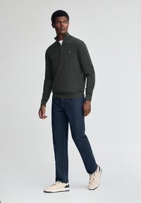 Mörkgrå pullovrare med halvdragkedja, texturerad tyg, kombinerad med blåjeans och ljusa sneakers, stående mot en neutral bakgrund.